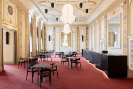 Heller Veranstaltungsraum mit aufwendig verzierter Stuckdecke in Weiß und Gold, rotem Teppichboden und mehreren runden Bistrotischen mit schwarzen Stühlen. An der rechten Seite befindet sich eine dunkle Bartheke. Eine große weiße Pendelleuchte hängt zentral von der Decke. Der Raum wirkt durch große Wandspiegel und hohe Fenster großzügig und repräsentativ. 