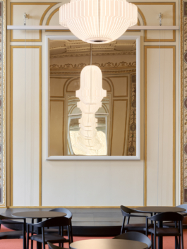 Heller Veranstaltungsraum mit aufwendig verzierter Stuckdecke in Weiß und Gold, rotem Teppichboden und mehreren runden Bistrotischen mit schwarzen Stühlen. An der rechten Seite befindet sich eine dunkle Bartheke. Eine große weiße Pendelleuchte hängt zentral von der Decke. Der Raum wirkt durch große Wandspiegel und hohe Fenster großzügig und repräsentativ. 