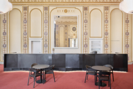 Heller Veranstaltungsraum mit aufwendig verzierter Stuckdecke in Weiß und Gold, rotem Teppichboden und mehreren runden Bistrotischen mit schwarzen Stühlen. An der rechten Seite befindet sich eine dunkle Bartheke. Eine große weiße Pendelleuchte hängt zentral von der Decke. Der Raum wirkt durch große Wandspiegel und hohe Fenster großzügig und repräsentativ. 