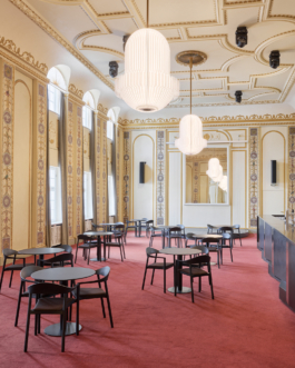 Heller Veranstaltungsraum mit aufwendig verzierter Stuckdecke in Weiß und Gold, rotem Teppichboden und mehreren runden Bistrotischen mit schwarzen Stühlen. An der rechten Seite befindet sich eine dunkle Bartheke. Eine große weiße Pendelleuchte hängt zentral von der Decke. Der Raum wirkt durch große Wandspiegel und hohe Fenster großzügig und repräsentativ. 