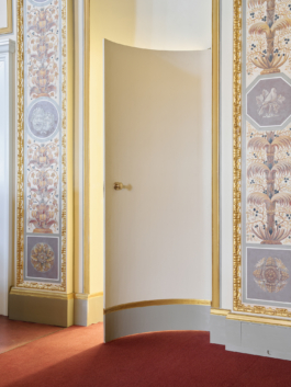 Heller Veranstaltungsraum mit aufwendig verzierter Stuckdecke in Weiß und Gold, rotem Teppichboden und mehreren runden Bistrotischen mit schwarzen Stühlen. An der rechten Seite befindet sich eine dunkle Bartheke. Eine große weiße Pendelleuchte hängt zentral von der Decke. Der Raum wirkt durch große Wandspiegel und hohe Fenster großzügig und repräsentativ. 