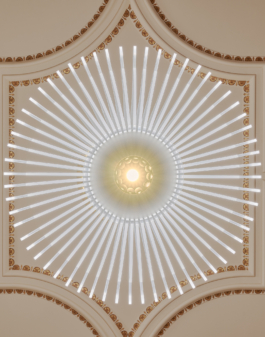 Heller Veranstaltungsraum mit aufwendig verzierter Stuckdecke in Weiß und Gold, rotem Teppichboden und mehreren runden Bistrotischen mit schwarzen Stühlen. An der rechten Seite befindet sich eine dunkle Bartheke. Eine große weiße Pendelleuchte hängt zentral von der Decke. Der Raum wirkt durch große Wandspiegel und hohe Fenster großzügig und repräsentativ. 