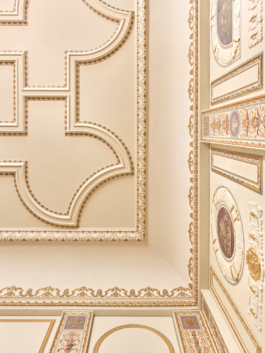 Heller Veranstaltungsraum mit aufwendig verzierter Stuckdecke in Weiß und Gold, rotem Teppichboden und mehreren runden Bistrotischen mit schwarzen Stühlen. An der rechten Seite befindet sich eine dunkle Bartheke. Eine große weiße Pendelleuchte hängt zentral von der Decke. Der Raum wirkt durch große Wandspiegel und hohe Fenster großzügig und repräsentativ. 