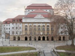 Heller Veranstaltungsraum mit aufwendig verzierter Stuckdecke in Weiß und Gold, rotem Teppichboden und mehreren runden Bistrotischen mit schwarzen Stühlen. An der rechten Seite befindet sich eine dunkle Bartheke. Eine große weiße Pendelleuchte hängt zentral von der Decke. Der Raum wirkt durch große Wandspiegel und hohe Fenster großzügig und repräsentativ. 