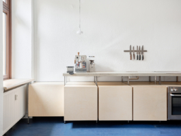 Schlichte, helle Küche mit blauem Boden: Entlang der rechten Wand stehen minimalistische Unterbauschränke aus hellem Holz mit einer durchgehenden Arbeitsplatte, darauf eine Siebträgermaschine; an der weißen Wand hängt eine magnetische Messerleiste, links fällt Tageslicht durch ein großes Fenster. 